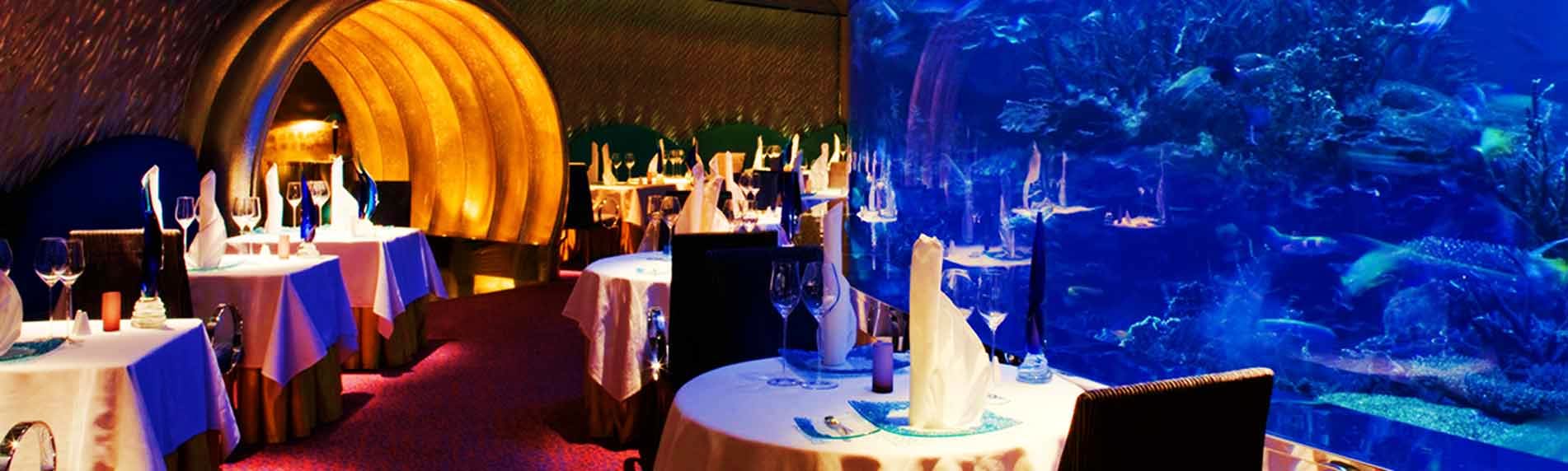 Underwater fine dining restaurant with large aquarium window displaying tropical fish and coral reef, elegant white tableclot
